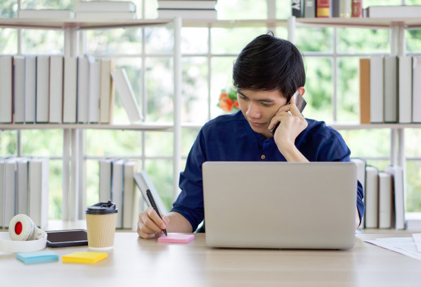 年轻的亚洲人做笔记，同时听手机。在家工作的概念。图片下载