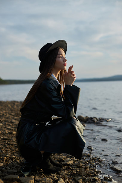 为日落时的年轻女子画像。穿着黑色圆帽和皮革雨衣的时尚女子在湖边。美丽的妆容和明亮的红色唇膏在嘴唇上图片下载