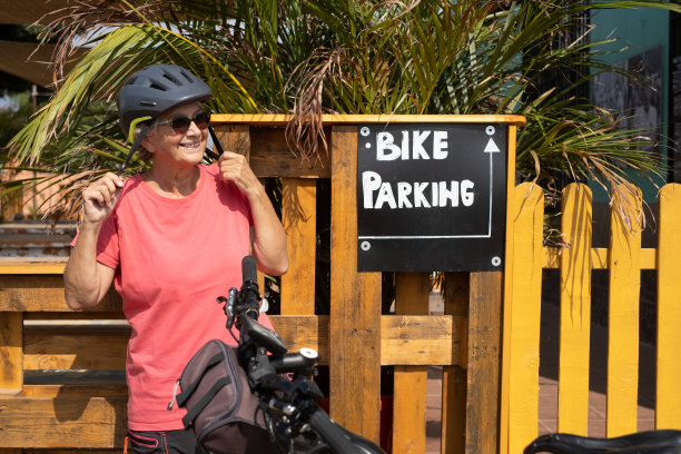 一位迷人的老妇人戴着防护头盔骑着自行车出游，停在有自行车停车场的餐前酒吧图片下载