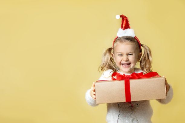 送圣诞礼物。微笑快乐兴奋的小女孩戴着红色圣诞帽摆出孤立的黄色背景拿着牛皮纸纸盒。网上购物，个人产品销售。积极的脸。模拟、空间文本图片下载