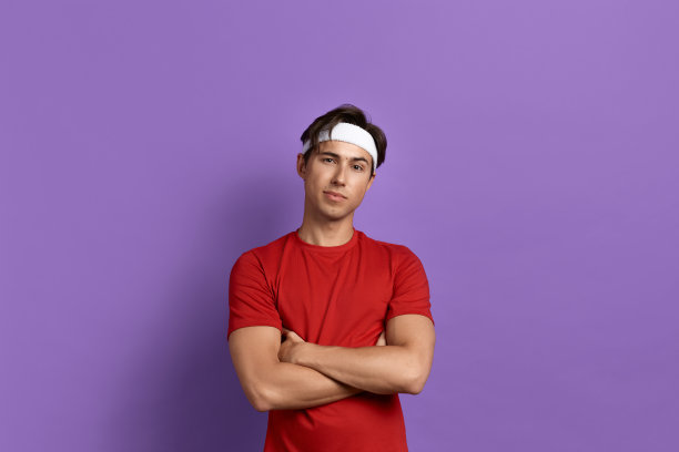 友好的年轻运动员或教练双臂交叉在胸前，是健身成瘾，提倡健康的生活方式，正在去工作，穿着红色的t恤和白色的头带，孤立在紫色的背景。图片下载