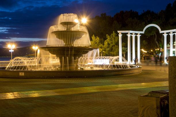 歌唱三层喷泉和拱门与柱子的背景上的日落天空。傍晚葛伦芝克路堤。路堤上有栏杆，灯光闪烁。喷泉和拱门后面的树木剪影图片下载