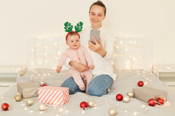快乐乐观的女妈妈，带着节日的心情，抱着宝宝，拿着手机，和她可爱可爱的蹒跚学步的女儿拍照。图片下载