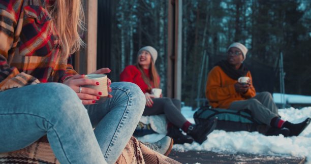 特写女性手握热饮杯，快乐的朋友坐在豆袋上聊天在温暖的冬季阳台慢动作。图片下载
