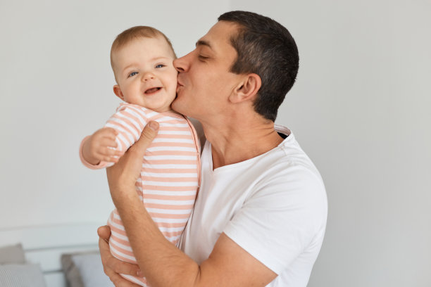 穿着白色休闲t恤的快乐黑发男人抱着他的女儿，亲吻她的脸颊，表达了伟大的生活，在家里的光室摆姿势。图片下载