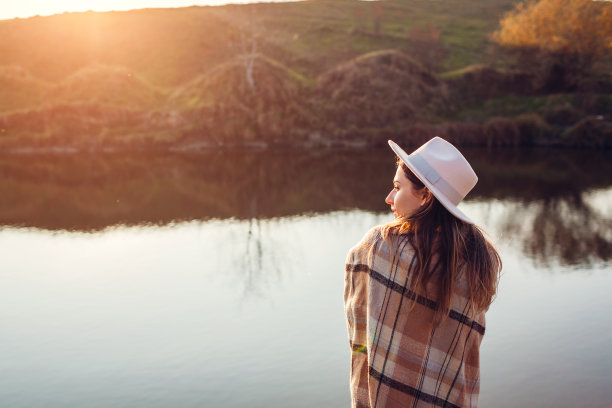 后视图的女人放松在秋天的河流在日落。戴着帽子的时尚女孩静静地享受着秋天的风景图片下载