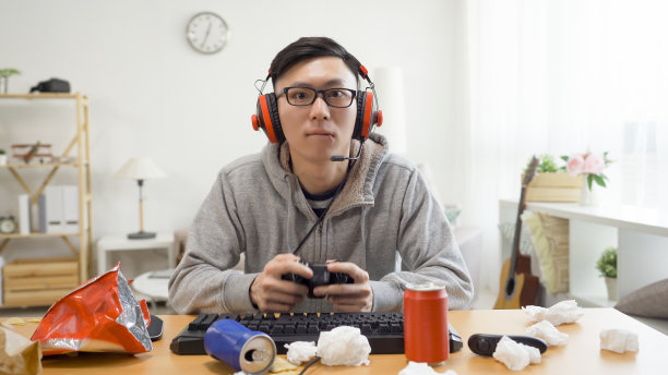 亚洲十几岁的男孩玩视频游戏，而放松在家从学校的暑假。戴着眼镜戴着耳机的近视眼男子拿着快乐棒在肮脏的桌子上玩，室内有垃圾食品。图片下载