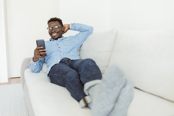 一个年轻的美国黑人，穿着牛仔休闲服，坐在舒适的米色沙发上，用他的智能手机浏览网页。图片下载