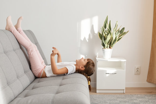 侧视图肖像的小女孩穿着白色t恤躺在沙发上抬起双腿，感到无聊，不知道做什么，独自在家里度过学校假期。图片下载