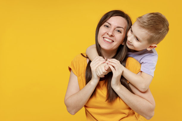 快乐的年轻女人有有趣的可爱的婴儿男孩5-6-7岁，穿着紫色t恤。在黄色背景的演播室里，妈妈、小孩、儿子一起摆姿势，互相拥抱。母亲节爱家庭观念。图片下载