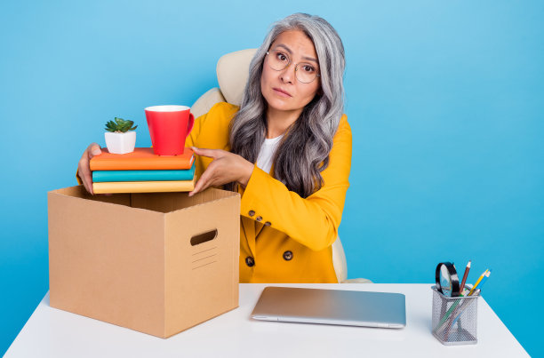 照片中不快乐抑郁的成熟女性穿着黄色夹克眼镜，包装东西失去工作孤立的蓝色背景图片下载