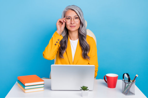 照片上漂亮自信的高级女士穿着黄色西装，戴着眼镜微笑着写着现代小玩意，工作场所孤立的蓝色背景图片下载