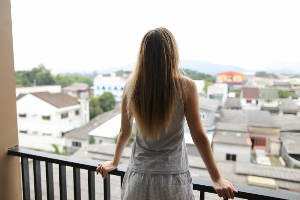 小女孩穿着夏天的睡衣站在阳台上，看着背景中的建筑物图片下载