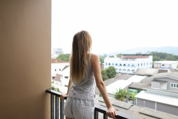 年轻的金发女孩穿着夏天的睡衣站在阳台上，看着背景中的建筑物图片下载