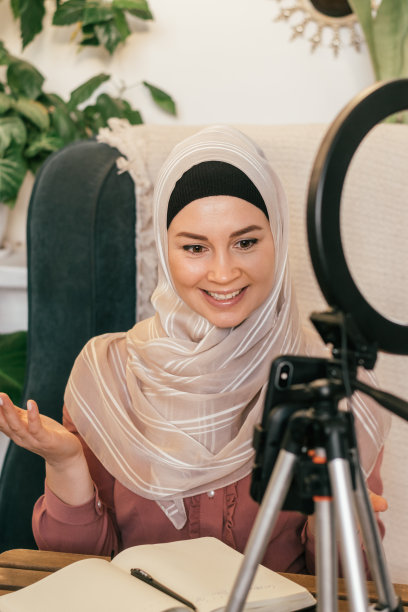 一位戴着头巾的年轻穆斯林妇女主持她的视频博客，并进行现场直播。舒适的室内室内植物。社交媒体和技术概念，多样化的人。图片下载