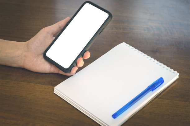 在木桌的背景上，一名女子拿着空白屏幕的手机。工作场所有记事本和笔。模型复制空间。图片下载