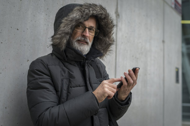一位头发花白、留着胡子的商人，穿着绿色冬季夹克，戴着兜帽，戴着眼镜，站在办公楼的混凝土墙旁边，正在打电话，盯着手机。图片下载
