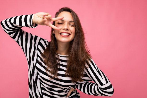 年轻的欧洲积极微笑迷人的快乐美丽的黑发女人与真诚的情绪穿着休闲条纹套衫孤立在粉红色的背景与复制空间图片下载