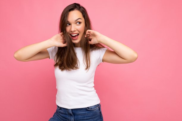 照片肖像年轻美丽的嬉皮士微笑黑发女人在白色t恤与模型。性感无忧无虑的女性在工作室的粉红色墙壁附近摆着孤立的空空间。自然妆容的正面模特图片下载