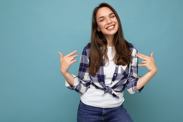 年轻的积极微笑美丽的黑发女人与真诚的情绪穿着时髦的格子衬衫孤立地站在蓝色的背景与自由的空间，并指向自己图片下载