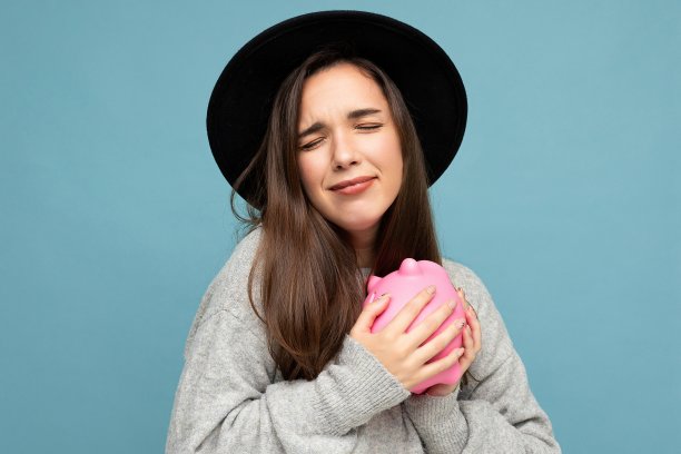 肖像照片的情绪积极敏感的年轻美丽有吸引力的黑发女人与真诚的情绪穿着灰色毛衣和黑色帽子孤立在蓝色的背景与复制空间和持有粉红色的小猪盒子。储钱罐的概念图片下载