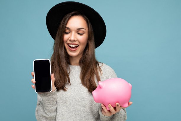 肖像照片的快乐积极微笑满意真诚年轻漂亮漂亮的好女人，黑头发穿着时尚的衣服和黑色的帽子孤立在蓝色的背景与复制空间，拿着粉红色的猪钱箱，显示手机与空屏幕图片下载