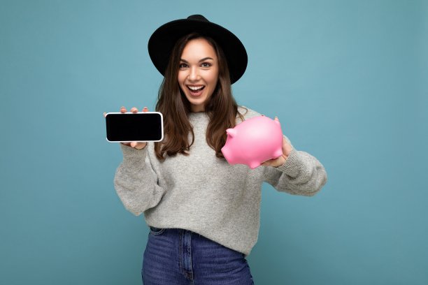 幸福正面微笑满意真诚年轻迷人的黑发女性人的肖像照片穿着时尚的灰色毛衣和黑色帽子孤立在蓝色背景与自由空间和持有粉红色猪便士银行和显示手机与空图片下载