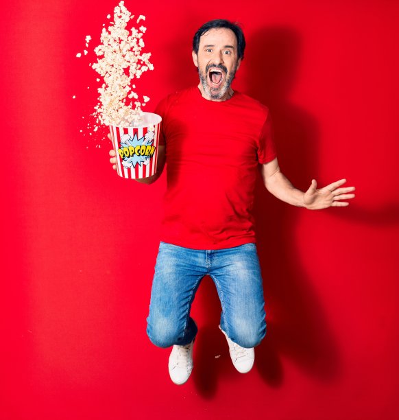 中年英俊的西班牙男子惊讶地张开嘴巴。拿着一桶爆米花跳过孤立的红色背景图片下载