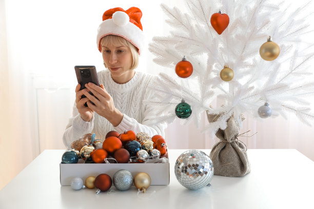 一个漂亮的女人装饰了一棵美丽的白色圣诞树，并在你的智能手机上自拍，假日，庆祝活动和人们的概念图片下载