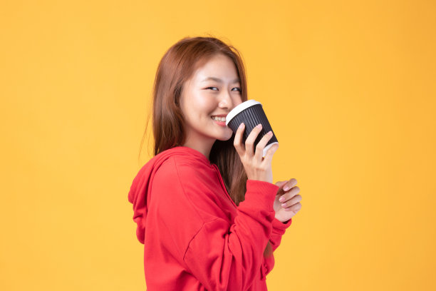 一个漂亮的年轻亚洲女人的肖像微笑，拿着外卖咖啡杯穿着红色毛衣孤立在黄色背景。美丽的年轻女孩喜欢早上喝咖啡。图片下载