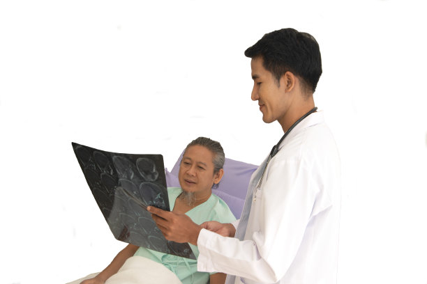 图为，亚洲男性医生正在用CT扫描患者的脑部x光片，并与躺在旁边白色背景下的患者讨论医疗问题。图片下载