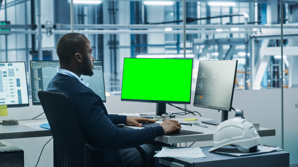 汽车工厂办公室:黑色工程师在电脑上工作，两个显示器屏幕显示色度键绿色屏幕和自动化机器人手臂装配线的监控制造高科技电动汽车图片下载