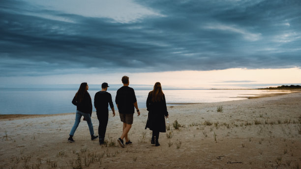 四个朋友在荒芜的海滩上散步。黄昏时平静的大海。他们离开镜头。一个女孩拿着酒杯。与大自然共度夏日时光。图片下载