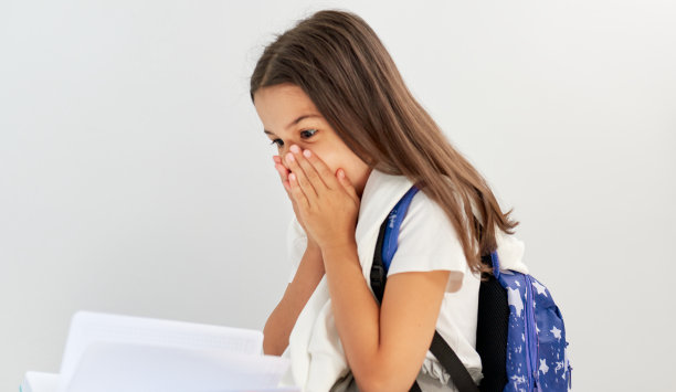 工作室图片:一个小女孩，穿着白色t恤，背着蓝色背包，坐在地板上读故事。当她得了最高分时，一个女学生露出惊讶的表情。图片下载