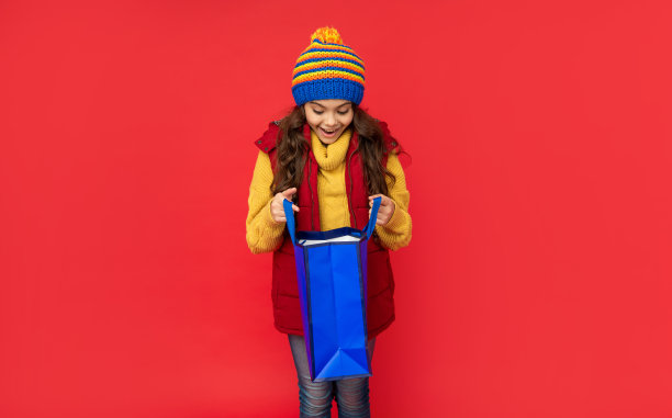 惊奇的孩子在冬天的衣服打开购物袋上的红色背景，圣诞节购物图片下载