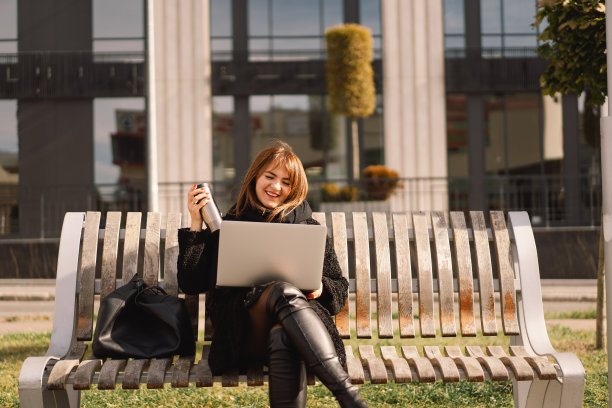 一个女人坐在城市的长椅上，一边用电脑一边喝茶。自由的女人。图片下载
