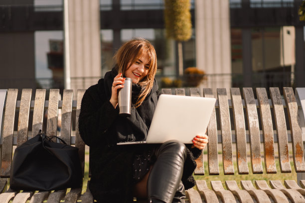 一个女人坐在城市的长椅上，一边用电脑一边喝茶。自由的女人。图片下载
