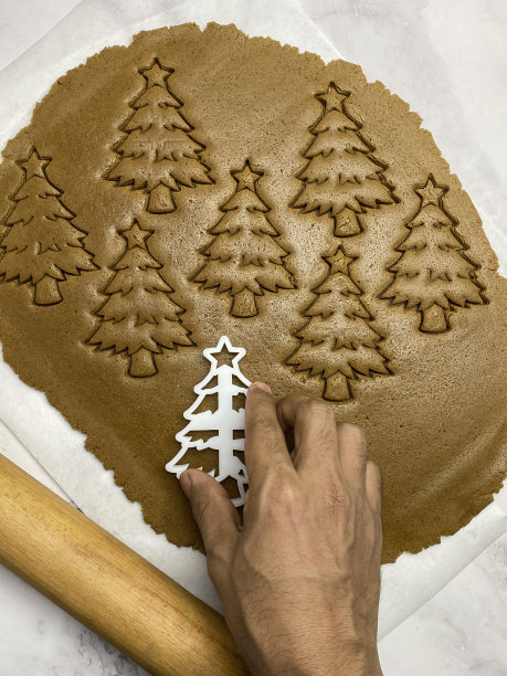 照片中，一个不认识的人正在用塑料圣诞树饼干刀切自制的饼干，姜饼曲奇面团滚在防油的羊皮纸上，擀面杖，树的形状，家庭烘焙，高架视图图片下载