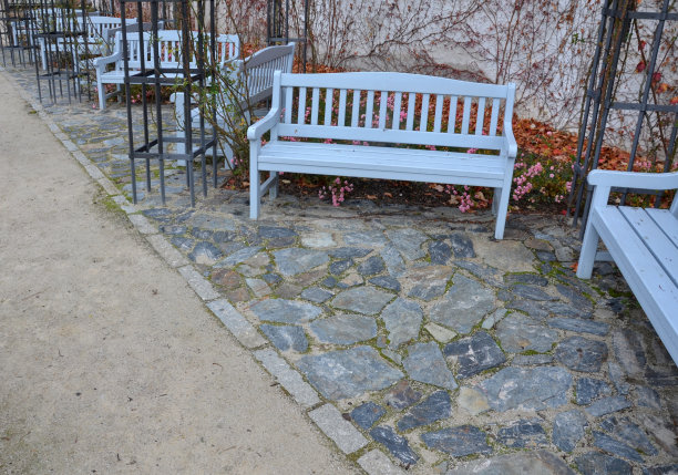 公园里的石质灰色路面上有木凳，低矮的挡土墙堆积着斑岩。玫瑰园的玫瑰园。地被，树和攀缘玫瑰在玫瑰与锻造金属图片下载