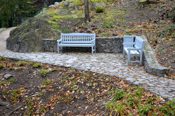 公园里的石质灰色路面上有木凳，低矮的挡土墙堆积着斑岩。玫瑰园的玫瑰园。地被，树和攀缘玫瑰在玫瑰与锻造金属图片下载