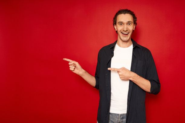 年轻开朗又惊讶又困惑的男学生穿着休闲的蓝色衬衫，用食指指着旁边自由工作区的模拟复制空间孤立在红色背景的工作室肖像。促销活动图片下载