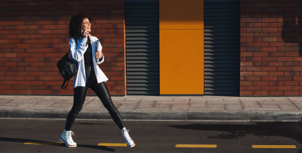 一个女人走在城市的街道上打电话。从一个手持笔记本电脑和平板电脑的学生的侧面看。砖墙。阳光灿烂的日子图片下载