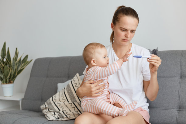 室内拍摄的年轻母亲抱着她蹒跚学步的婴儿坐在床上，拿着妊娠测试结果呈阳性的照片，不知道如何应对意外怀孕。图片下载