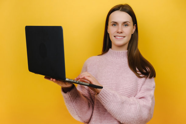 微笑可爱的广告撰稿人自由职业者年轻的白种人女人的肖像手持笔记本电脑聊天打字文字，快乐的看着相机，摆出孤立的黄色背景墙在工作室图片下载