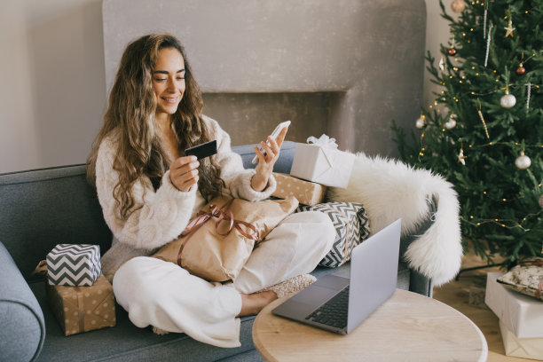 一名年轻女子在装饰着圣诞树和节日彩灯的舒适房间里用智能手机、笔记本电脑和信用卡在线购物。图片下载