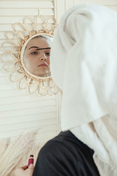 年轻漂亮的女人在浴室淋浴后用毛巾蒙着头画睫毛，看着墙上镜子里自己的倒影。女性早上化妆程序的概念。图片下载