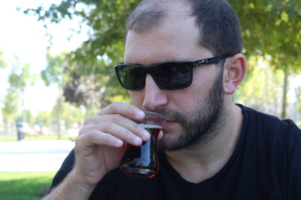 留着胡子的黑发男人戴着墨镜，坐在绿色的草地上喝着玻璃杯里的红茶图片下载