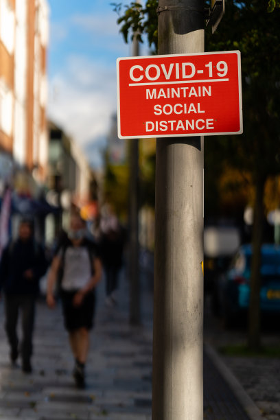 灯柱上红色和白色的“COVID-19保持社交距离”标志，背景中有视线模糊的行人。副本的空间。图片下载
