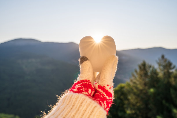 女性的手在圣诞手套抱雪在一个形状的心在山的背景与阳光。图片下载