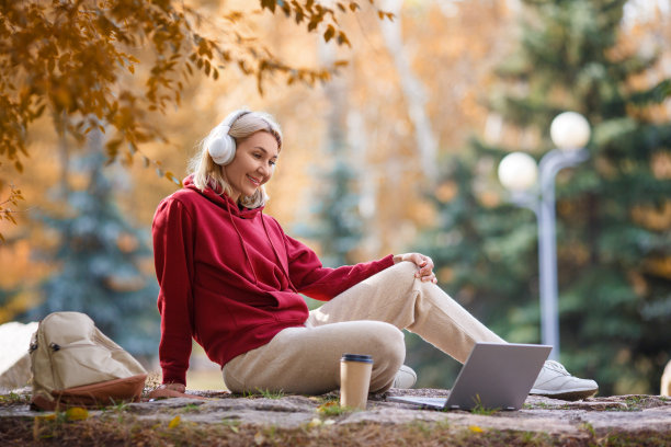 在线会议。秋天的户外公园里，一位穿着便服、头戴耳机的友好女士坐在笔记本电脑前，喝着咖啡。图片下载